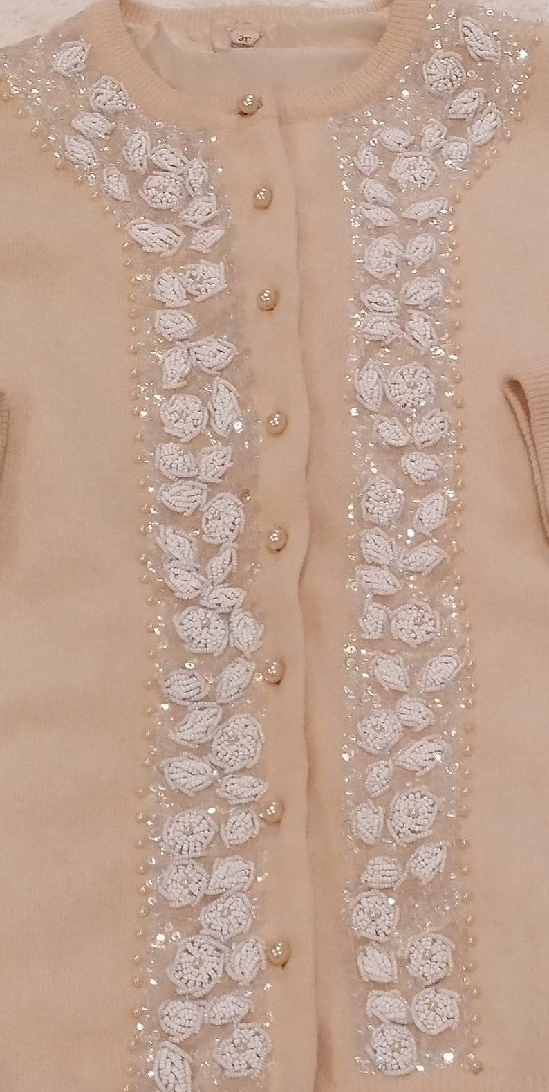 Op de afbeelding: Een beige cardigan met witte parelmoer bloemenborduursel aan de voorkant. De cardigan heeft een knoopsluiting.