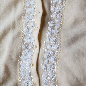Op de afbeelding: Een cr&egrave;mekleurige cardigan met witte parelversieringen aan de voorkant. De cardigan heeft een knoopsluiting en een geribbelde zoom.