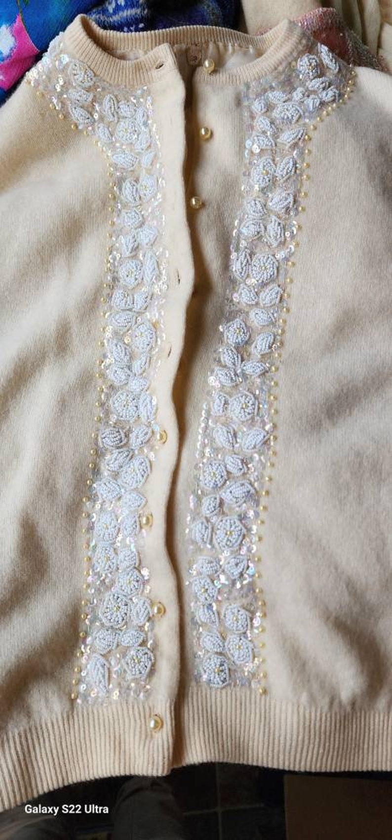 Op de afbeelding: Een cr&egrave;mekleurige cardigan met witte parelversieringen aan de voorkant. De cardigan heeft een knoopsluiting en een geribbelde zoom.