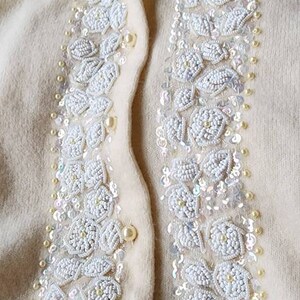 Op de afbeelding: Een cr&egrave;mekleurige cardigan met witte parelmoer-bloemenborduursel aan de voorkant. De cardigan heeft een knoopsluiting en een geribbelde zoom.