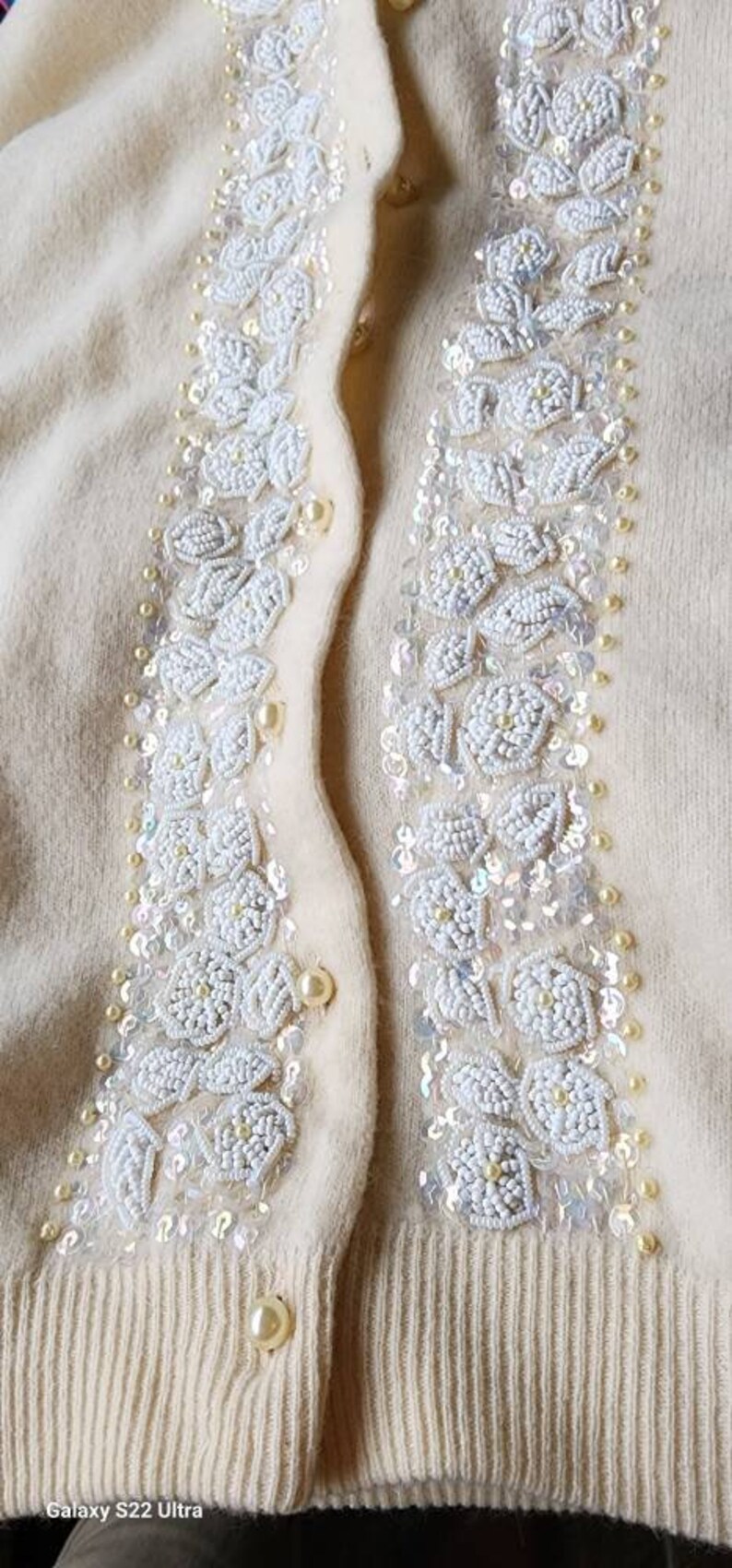 Op de afbeelding: Een cr&egrave;mekleurige cardigan met witte parelmoer-bloemenborduursel aan de voorkant. De cardigan heeft een knoopsluiting en een geribbelde zoom.