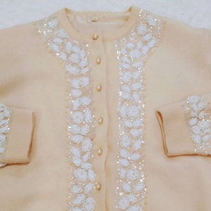 Op de afbeelding: Een cr&egrave;mekleurige cardigan met witte parelversieringen aan de voorkant en mouwen. De cardigan heeft een knoopsluiting.