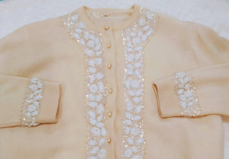 Op de afbeelding: Een cr&egrave;mekleurige cardigan met witte parelversieringen aan de voorkant en mouwen. De cardigan heeft een knoopsluiting.