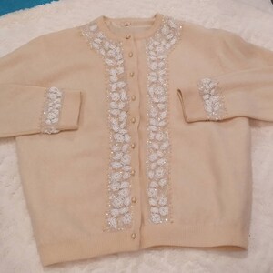 Op de afbeelding: Een cr&egrave;mekleurige cardigan met witte parelversieringen aan de voorkant en manchetten. De cardigan heeft een knoopsluiting.