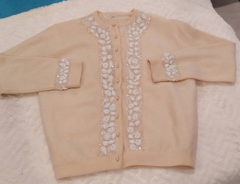 Op de afbeelding: Een cr&egrave;mekleurige cardigan met witte parelversieringen aan de voorkant en manchetten. De cardigan heeft een knoopsluiting.