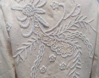 Puede incluir: Un cárdigan de color crema con un diseño floral bordado con cuentas blancas. El diseño presenta una flor grande con hojas y flores más pequeñas.