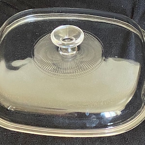 Op de afbeelding: Doorzichtige glazen deksel met een ronde knop. De deksel heeft een reliëf, getextureerd patroon rond de knop.