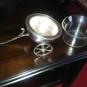 May include: A silver cart with a single wheel and a silver bowl on top. The cart has a handle and is sitting on a dark wooden surface. Next to the cart is a clear glass bowl.