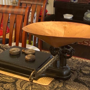 May include: Antique black and copper scale with a large, round, copper bowl. The scale has a black platform with three round weights on it. The scale is sitting on a patterned table cloth.