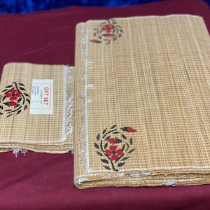 May include: A set of two brown woven placemats with white fringe and red floral designs. The placemats are labeled "Gift Set".