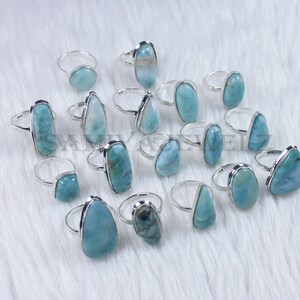 May include: A collection of 15 silver rings with blue and green gemstone settings. The rings are arranged on a white fuzzy surface.