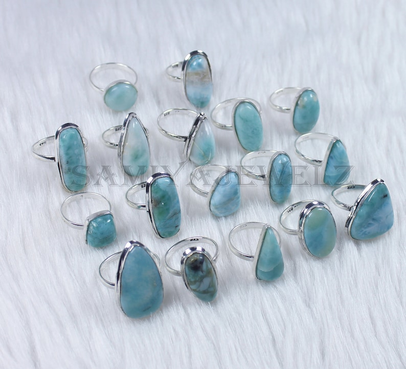 May include: A collection of 15 silver rings with blue and green gemstone settings. The rings are arranged on a white fuzzy surface.