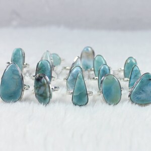 May include: A collection of 14 silver rings with blue and green gemstone settings. The rings are arranged in a row on a white fuzzy surface.