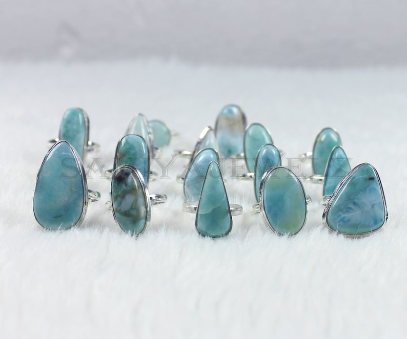 May include: A collection of 14 silver rings with blue and green gemstone settings. The rings are arranged in a row on a white fuzzy surface.