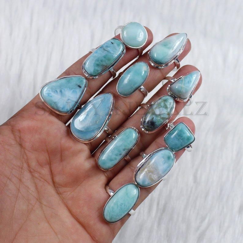 May include: A collection of 12 rings featuring blue and green gemstone cabochons set in silver. The rings are displayed on a hand.