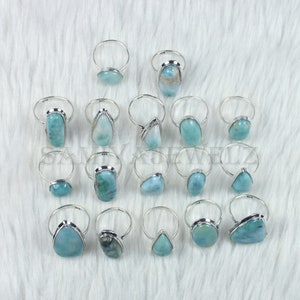 May include: A collection of 16 silver rings with blue and green gemstone settings. The rings are arranged in a semi-circle on a white fuzzy surface.