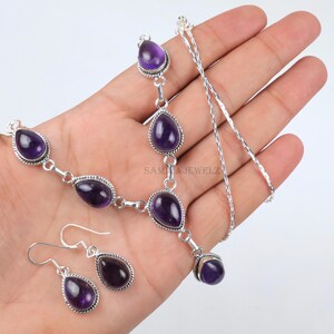 May include: A silver necklace and earring set featuring teardrop-shaped, deep purple amethyst stones. The necklace has multiple stones connected by silver links, and the earrings have a single stone each. The jewelry is displayed on a light background.