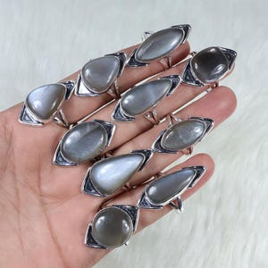 Puede incluir: Una colección de anillos de plata con cabujones de piedra lunar gris de diversas formas, como ovaladas y en forma de lágrima. Los anillos se muestran en una mano sobre un fondo blanco y esponjoso. Cada anillo tiene un diseño único.