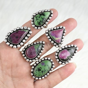 Puede incluir: Un conjunto de anillos con piedras preciosas y engastes plateados. Los anillos presentan diversas formas, como óvalos, lágrimas y triángulos. Las piedras preciosas muestran tonos verdes y rojos, con un aspecto texturizado. Los anillos se muestran en una mano.
