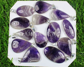 May include: A collection of eleven polished amethyst pendants in various oval and teardrop shapes, each set in a silver-toned metal frame with a bail. The stones feature deep purple and white banded patterns, arranged on a white display card.