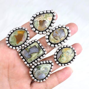 May include: A collection of silver-toned rings with various gemstone shapes, including oval, teardrop, and rectangular. The gemstones feature green, brown, and gray hues, set in a beaded border.