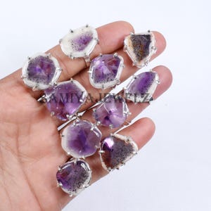 以下が含まれることがあります： 不規則な形状のアメジスト宝石をあしらったシルバーリングのコレクション。アメジストは、白と茶色のインクルージョンを伴うさまざまな紫の色合いを示しています。リングは明るい背景に表示されています。