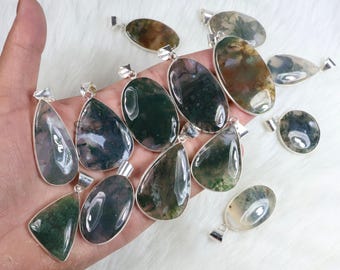 May include: A collection of moss agate pendants in various shapes, including oval, teardrop, and triangular. Each pendant features a unique pattern of green and brown inclusions set in a silver-colored bezel. The pendants are displayed on a white, fluffy surface.