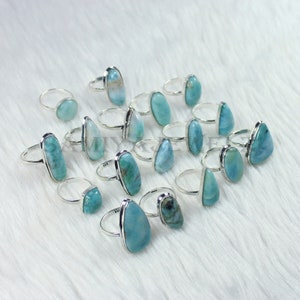 May include: A collection of 15 silver rings with blue and green gemstone settings. The rings are arranged on a white fuzzy surface.