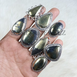 May include: A collection of silver-toned rings with various shapes, each featuring a polished, golden-colored stone. The rings are displayed on a light-colored, textured surface. The stones have a metallic sheen, and the settings have intricate detailing.