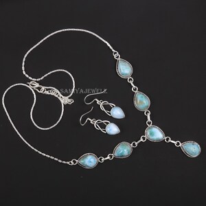 K&ouml;nnte beinhalten: Ein silbernes Halsketten- und Ohrring-Set. Die Halskette und die Ohrringe weisen tropfenf&ouml;rmige, hellblaue Edelsteine auf. Der Schmuck wird auf schwarzem Hintergrund pr&auml;sentiert. Der Markenname "SAMYAJEWELZ" ist sichtbar.