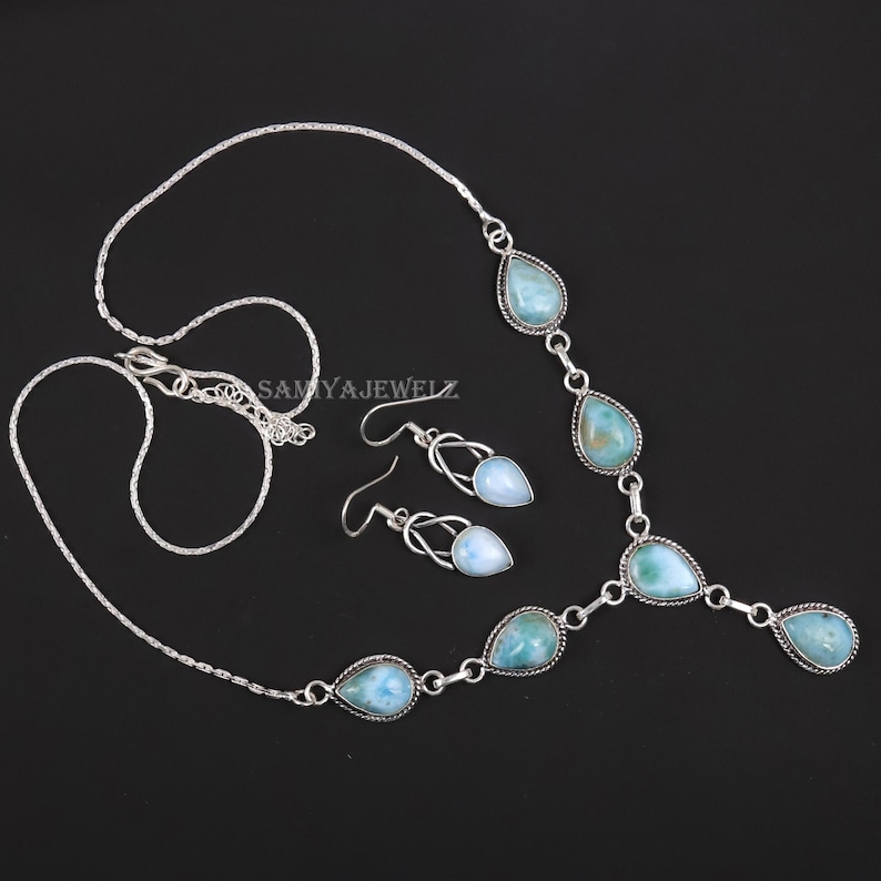 K&ouml;nnte beinhalten: Ein silbernes Halsketten- und Ohrring-Set. Die Halskette und die Ohrringe weisen tropfenf&ouml;rmige, hellblaue Edelsteine auf. Der Schmuck wird auf schwarzem Hintergrund pr&auml;sentiert. Der Markenname "SAMYAJEWELZ" ist sichtbar.