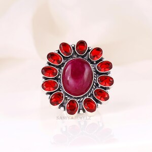May include: A silver ring featuring a large oval, deep red gemstone at the center, surrounded by a halo of smaller, oval red gemstones. The ring's design is floral-inspired, with the text "SAMIYAJEWELZ" visible.