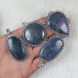 May include: Four gemstone pendants in various shapes: oval, round, teardrop, and oblong. Each pendant features a blue-green stone with red flecks, set in a silver-colored, beaded frame. The pendants are displayed on a white, textured surface.