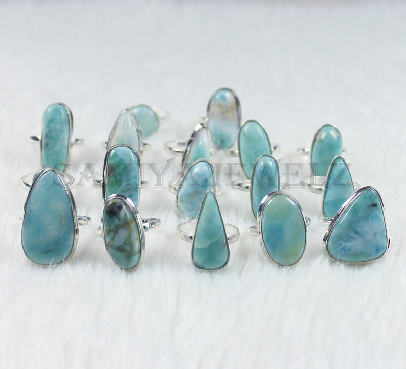 May include: A collection of 15 silver rings with blue and white gemstone settings. The rings are arranged on a white fuzzy surface.
