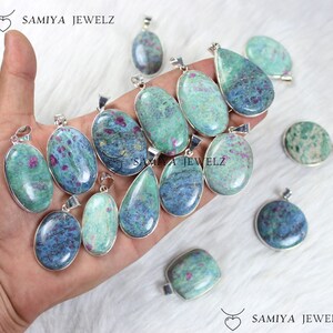 Può includere: Collezione di pendenti con pietre preziose di varie forme, tra cui ovali, a goccia e rotondi. I pendenti presentano una miscela di tonalità blu e verdi con inclusioni rosse, incastonati in metallo color argento. Il nome del marchio "SAMIYA JEWELZ" è visibile.