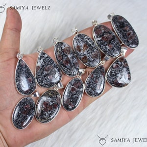 Peut inclure: Plusieurs pendentifs en pierres précieuses de formes variées, dont ovales et en goutte. Les pendentifs présentent un motif moucheté de teintes noires, blanches et rouge-brun, sertis dans du métal argenté. La marque "SAMIYA JEWELZ" est visible.