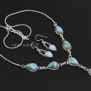 K&ouml;nnte beinhalten: Ein silbernes Halsketten- und Ohrring-Set mit tropfenf&ouml;rmigen, hellblauen Edelsteinen. Die Halskette und die Ohrringe haben ein filigranes Design. Der Schmuck wird vor einem schwarzen Hintergrund pr&auml;sentiert.