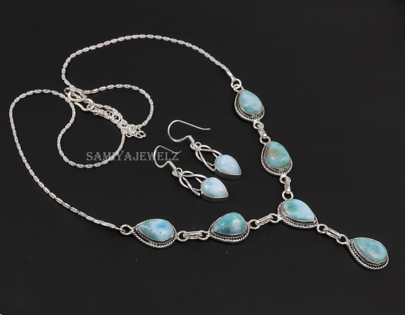 K&ouml;nnte beinhalten: Ein silbernes Halsketten- und Ohrring-Set mit tropfenf&ouml;rmigen, hellblauen Edelsteinen. Die Halskette und die Ohrringe haben ein filigranes Design. Der Schmuck wird vor einem schwarzen Hintergrund pr&auml;sentiert.