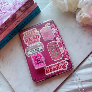 May include: A pink tablet case covered in stickers with phrases like "Hot Girls Read Books" and "Aggressively Reading." The case is next to a stack of books and white flowers.