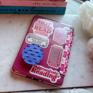Op de afbeelding: Een roze tablet hoesje versierd met stickers met teksten als "Hot Girls Read Books" en "Aggressively Reading." Daarnaast een stapel boeken met titels als "So Wrong" en "Lights Out." Het hoesje heeft een patroon.