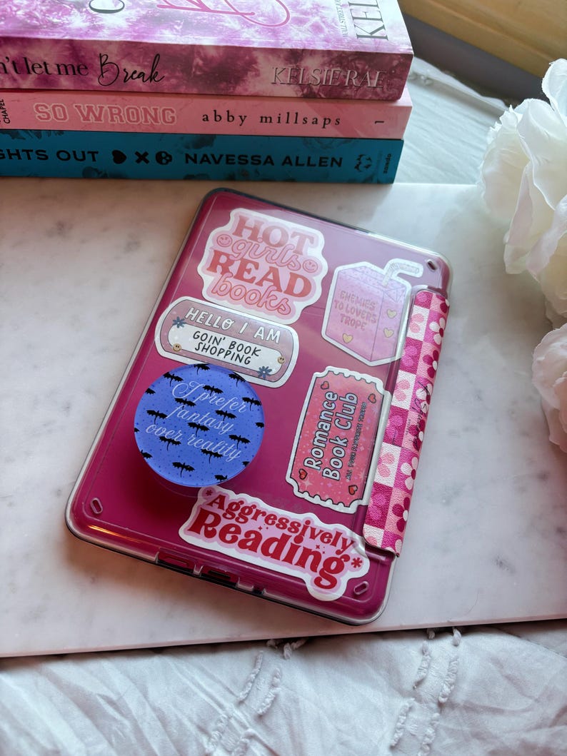 Op de afbeelding: Een roze tablet hoesje versierd met stickers met teksten als "Hot Girls Read Books" en "Aggressively Reading." Daarnaast een stapel boeken met titels als "So Wrong" en "Lights Out." Het hoesje heeft een patroon.