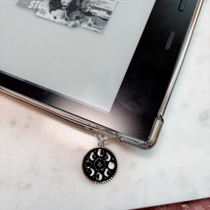 May include: A black and white moon phases charm attached to a clear plastic book cover. The charm is hanging from a silver chain.