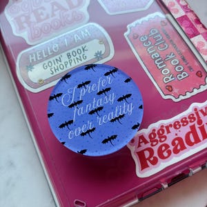 Op de afbeelding: Een blauwe telefoonhouder met de tekst "I prefer fantasy over reality" en zwarte vleermuisillustraties. De houder bevindt zich op een roze telefoonhoesje bedekt met stickers met boeken thema's.