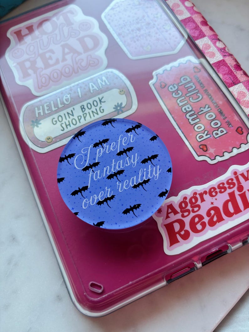 Op de afbeelding: Een blauwe telefoonhouder met de tekst "I prefer fantasy over reality" en zwarte vleermuisillustraties. De houder bevindt zich op een roze telefoonhoesje bedekt met stickers met boeken thema's.