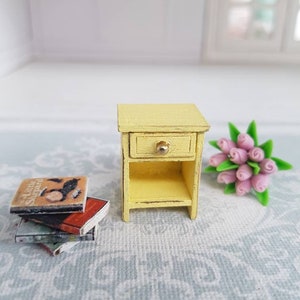 May include: A miniature yellow wooden nightstand with a single drawer and a shelf. The drawer has a gold knob. The nightstand is on a light gray patterned surface with a stack of miniature books and a bouquet of pink roses.