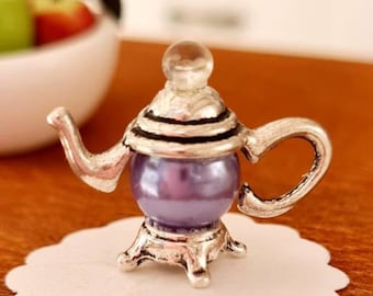 May include: A miniature silver teapot with a purple glass body and a clear glass knob on the lid. The teapot is sitting on a white scalloped doily.