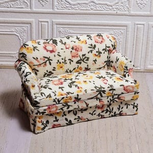 May include: A miniature sofa with a floral pattern in pink, yellow, and green on a cream background. The small couch has a rounded back and armrests, and is designed for a dollhouse or similar setting.