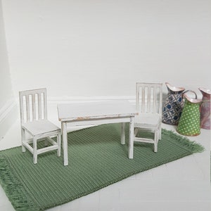 May include: A white miniature table and two chairs set on a green rug. The table and chairs are distressed and have a whitewashed finish. The set is displayed in a dollhouse setting with signs that read "Farm Fresh Eggs" and "Free Range Eggs" hanging on the wall.
