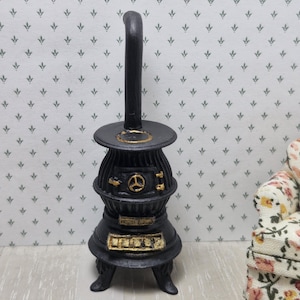 Puede incluir: Una estufa negra en miniatura con detalles dorados, que incluye una chimenea alta y elementos decorativos. La estufa tiene una parte superior redonda y una pequeña puerta con un diseño circular. Una pieza encantadora para una casa de muñecas o una escena en miniatura.