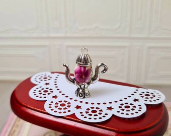 May include: A miniature silver teapot charm with a floral design on a white doily on a red table.